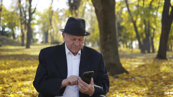 Thoughtful Senior Man in Suit and Hat Sits on Bench and Uses Smartphone in Park