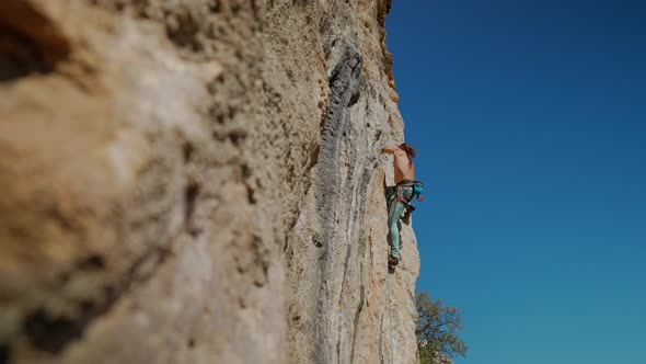 Bottom View of Muscular Strong Man Rock Climber Climbs on Vertical Cliff on Rock Wall on Blue Sky