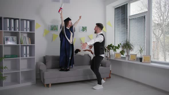 Young Man Hugs and Circles Happy Female Student in Academic Gown with Diploma in Hands Rejoice at