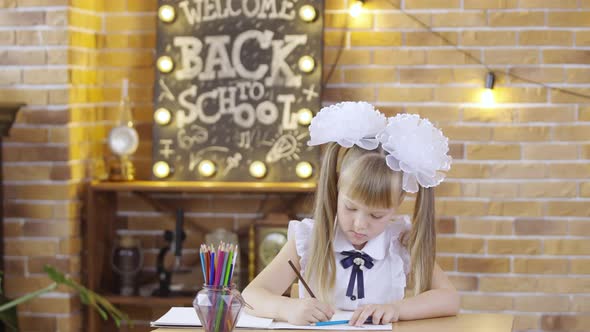 Cute Little Girl Sits at Table and Draws with Colored Pencils on the Background of a School Class