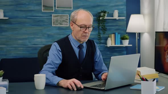 Tired Elderly Man Sitting at Desk Drinking Coffee Reading and Typing at Laptop