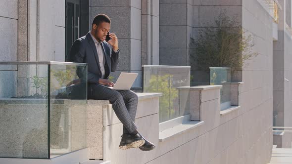 Young Adult Confident African American Businessman Using Laptop Outdoors Holding Smartphone Business