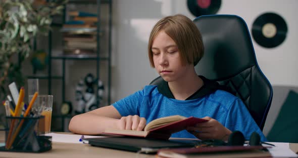 A Young Boy is Sitting at Desk Reading His Favorite Book