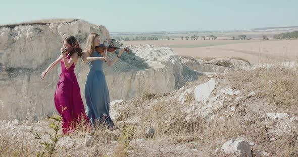 Virtuoso Violinists in Elegant Blowing Dresses Play the Music Among Rocks