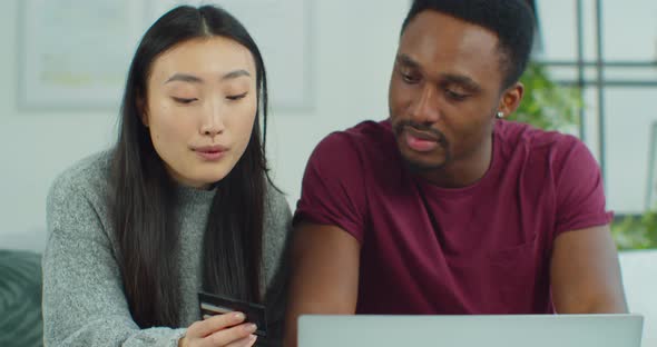 Cheerful Young Mixed Ethnicity Couple Shopping Online While Sitting Home with Credit Card and Laptop