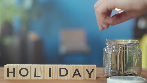 Hand Puts Money in Jar Next to Which There are Cubes with Word Holiday