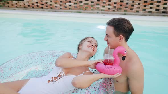 Young woman on inflatable ring gives to drink cocktail to her boyfriend into a swimming pool