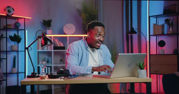 African American Sitting at the Desk in front of Laptop in Night Lighting Room