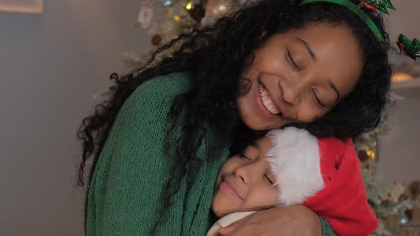 Happy Mom Hugs and Kisses Her Little Daughter in a Christmas Hat Near a Festive Tree