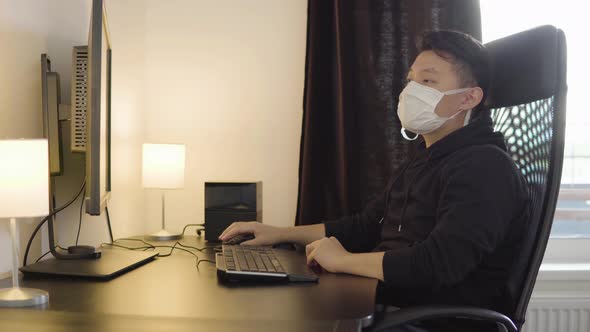 A Young Asian Man in a Face Mask Works on a Desktop Computer at Home, Tired