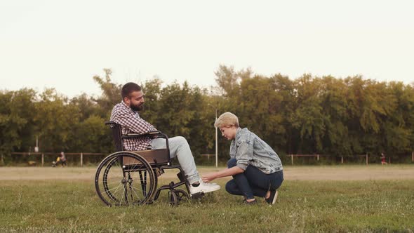 Loving couples ties his shoelaces to disabled person man sitting in wheelchair on grass in park