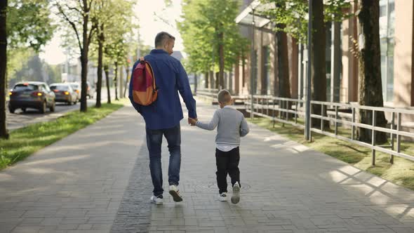 Back View of the Father Carrying Backpack Holding Sons Hand and Son is Holding the Plant in a Pot