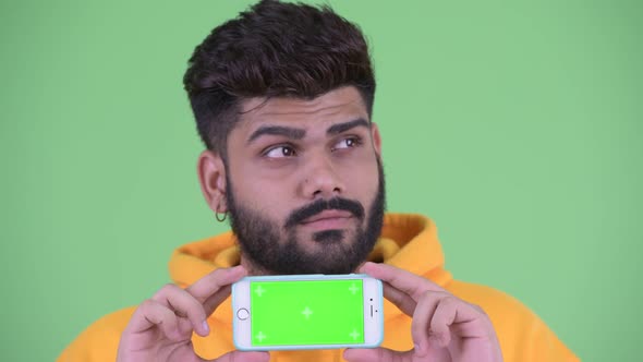 Face of Happy Young Overweight Bearded Indian Man Thinking While Showing Phone