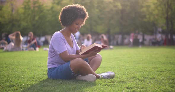 Smiling Young African Woman Reading Book Outdoor Relaxing on Lawn in Park