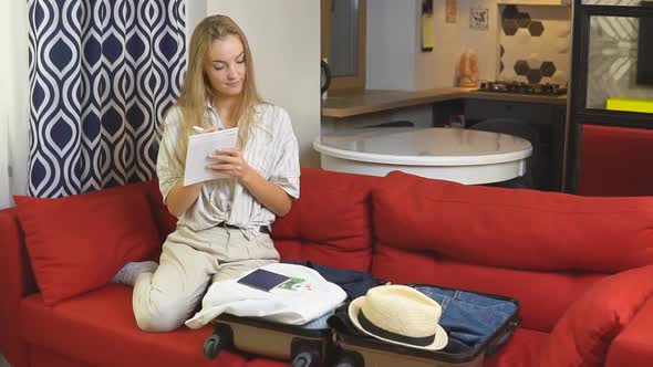 Young blonde woman packing suitcase making list of clothes.