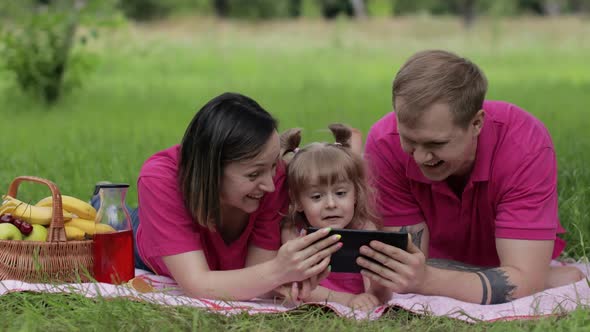 Family Weekend Picnic. Daughter Child Girl with Mother and Father Play Online Games on Tablet