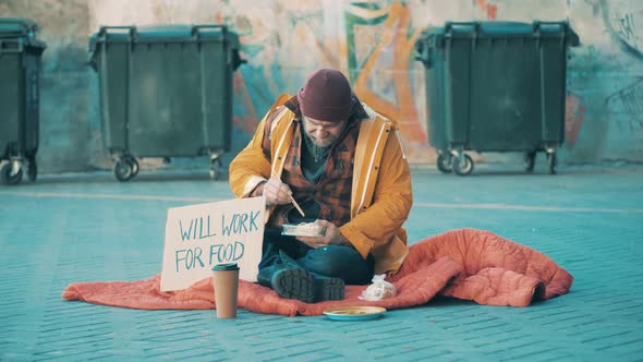 A Beggar is Eating on the Ground Near Waste Bins