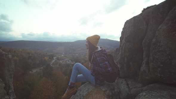 Girl Hiker in Checkered Jacket and Hat with Backpack Sits on Edge of Cliff and Enjoing Beautiful