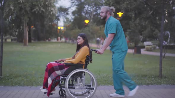 Side View Wide Shot of Volunteer Rolling Wheelchair with Handicapped Woman in Summer Park. Portrait