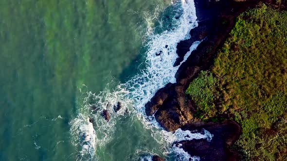Aerial shot turning above sunlit waves crashing into rocky California cliffs