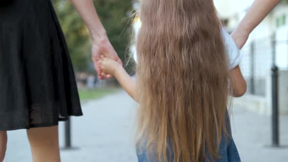 Back view of mother and her little daughter child walking together holding hands in summer park.