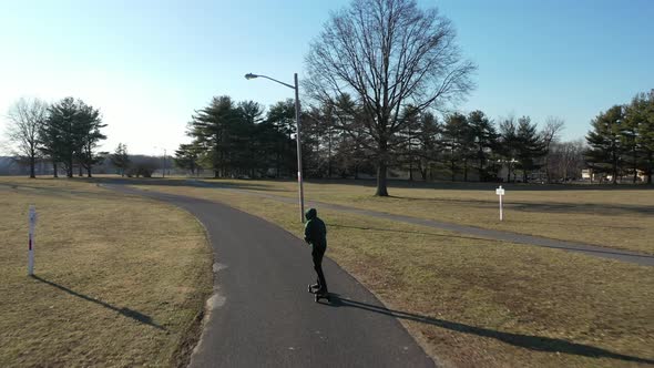 An aerial tracking of a man on an electric skateboard in an empty park on a sunny day. The drone fol