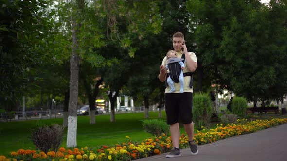 A Young Man Walks in the Park with a Newborn Child and Speaks on the Phone