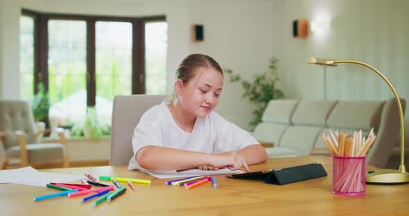 Focused Teen Girl at the Desk Scrolls Tablet Then Stretches Raising Hands Felttip Pens White Paper