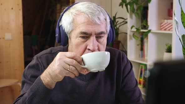 Senior Man in Headphones Seated at Table Looking at Laptop