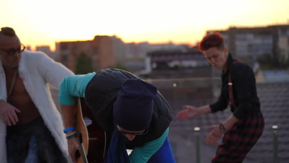 Joyful Adult Man Dancing with Guitar and Blurred Friends Having Fun at Background on Roof