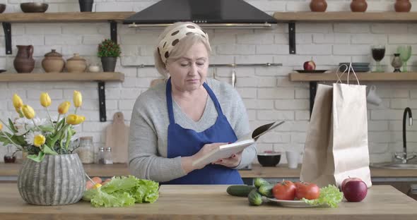 Positive Caucasian Senior Woman Looking for New Salad Recipe Standing in Kitchen