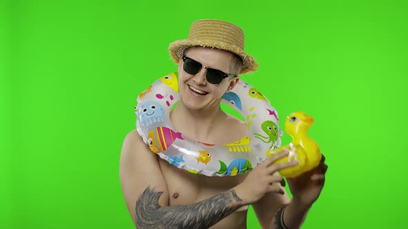 Shirtless Young Man Tourist with Swimming Ring on Shoulders Plays with Duck Toy