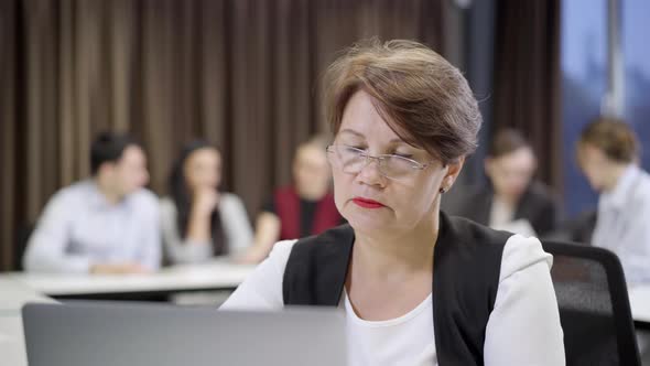 Concentrated Middle Aged Woman in Eyeglasses Surfing Internet on Laptop with Group of Blurred