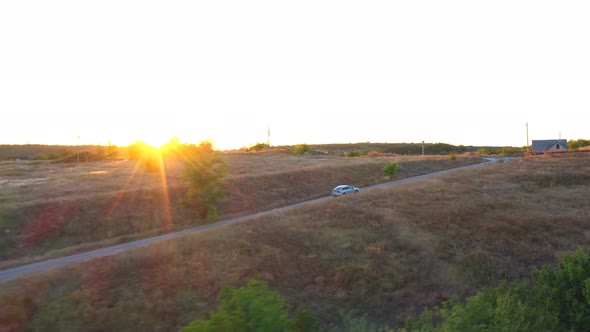 Aerial View of Electrical Car Driving on Country Road with Bright Sunset at Background