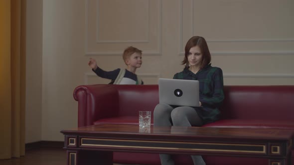 Young Working 30s Mom Sitting with Laptop on Home Sofa While Little Kid Coming Back From School with