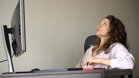 Tired Young Woman with Curly Hair and White Shirt is Working at the Office Using Her Computer