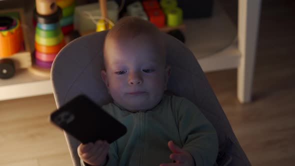 Cute Toddler Boy Sitting on Rocking Chair in Living Room and Watching Something on Mobile Phone