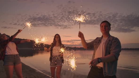 Cheerful Male and Female Friends are Running Along the Beach at Sunset Holding Sparkling Fireworks