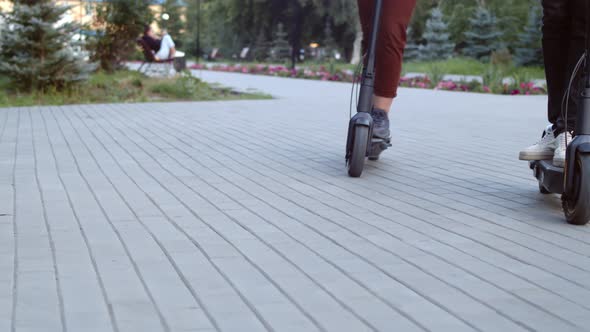 Male and Female Legs Ride on Electric Scooter in a Public Park Closeup Front View