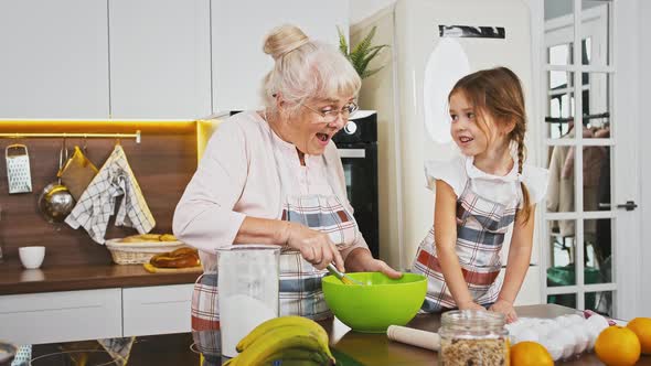 Granny Mixing Dough By Whisk in Bowl Little Granddaughter Putting Hands Into Flour and Clapping It