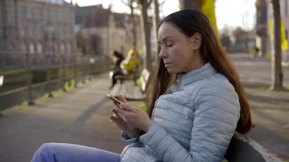 a Woman with Long Dark Hair and a Blue Jacket Sits on a City Bench on a Sunny Day. She Checks Her