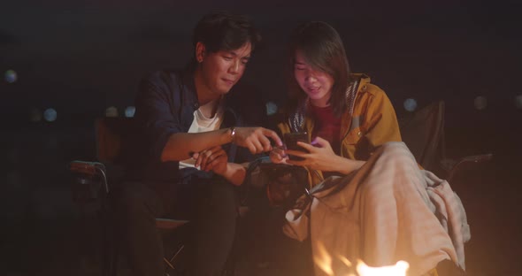 Young asia couple backpackers enjoy in camping at night near campfire on beach. using smart phone.