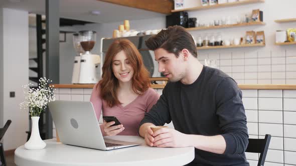 Two Young People Working with a Laptop and Smartphone in a Cafe and Drinking Coffee