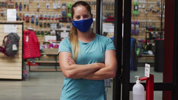 Caucasian female shopkeeper wearing face mask in the doorway of sports shop with arms crossed