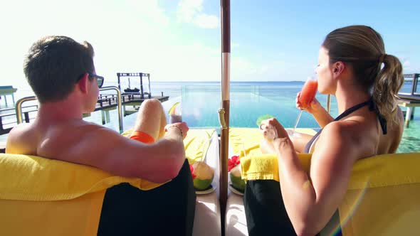 A couple relaxes and has drinks on an overwater bungalow at a tropical island resort hotel