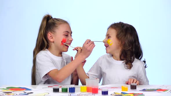 Two Little Girls Sit at the Table, Paint the Tassels on Their Faces, White Background, Slow Motion