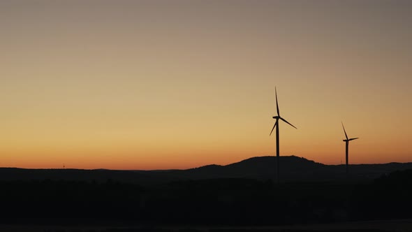 Static shot of the silhouette of two wind turbines spinning on a field at sunset