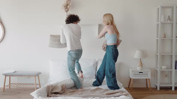 Curly Guy and Young Blonde Play Fighting With Pillows Standing On Bed in Modern Interior