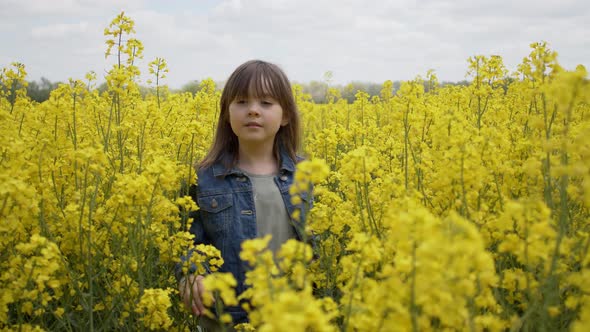 Smiling Young Girl in Jeans Jacket Walking Through Blooming Fresh Yellow Canola Field
