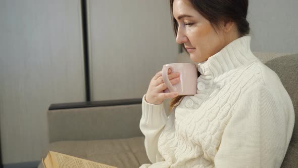 Young Woman in Sweater with Cup Tea Sitting on Sofa and and Reading a Book